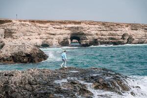 un mujer en un azul chaqueta soportes en un rock encima un acantilado encima el mar y mira a el furioso océano. niña viajero descansa, piensa, Sueños, disfruta naturaleza. paz y calma paisaje, Ventoso clima. foto