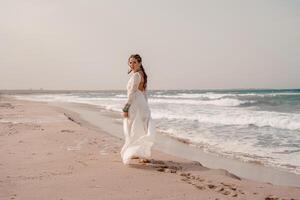 Model in boho style in a white long dress and silver jewelry on the beach. Her hair is braided, and there are many bracelets on her arms. photo