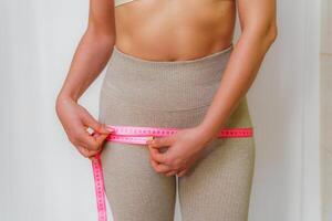 Cropped view of slim woman measuring hips with tape measure at home, close up. An unrecognizable European woman checks the result of a weight loss diet or liposuction indoors. Healthy lifestyle. photo