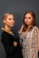 Two women are posing for a photo, one holding a hair curler. Scene is casual and friendly, as the women are smiling and looking at the camera. photo