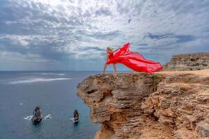 un mujer en un rojo seda vestir soportes por el océano, con montañas en el fondo, como su vestir se balancea en el brisa. foto