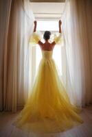 A woman's silhouette in a golden luxurious dress against the background of a window holds a curtain with her hands. Elegant lady in a yellow long silk dress with bare back, back view. photo