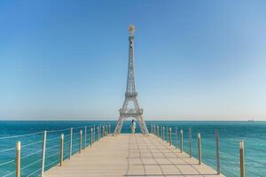 Large model of the Eiffel Tower on the beach. A woman walks along the pier towards the tower, wearing a blue jacket and white jeans. photo