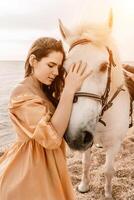 A woman in a dress stands next to a white horse on a beach, with the blue sky and sea in the background. photo