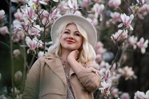 Magnolia flowers woman. A blonde woman wearing a hat stands in front of a tree with pink magnolia flowers. She is wearing a tan coat and a dress. photo