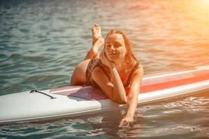 mujer cenar mar. Deportes niña en un tabla de surf en el mar en un soleado verano día. en un negro baños traje, él se sienta en un sapa en el mar. descanso en el mar. foto