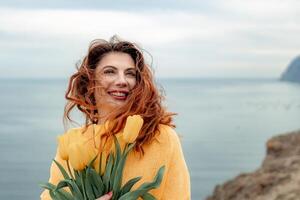 Portrait of a happy woman with hair flying in the wind against the backdrop of mountains and sea. Holding a bouquet of yellow tulips in her hands, wearing a yellow sweater photo