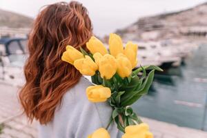 Woman holds yellow tulips in harbor with boats docked in the background., overcast day, yellow sweater, mountains photo