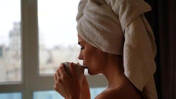 Middle aged woman looks good with bare shoulders in a white towel on her head holds a cup and drinks coffee or tea against the window photo