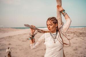 Model in boho style in a white long dress and silver jewelry on the beach. Her hair is braided, and there are many bracelets on her arms. photo