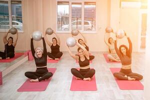 A group of six athletic women doing pilates or yoga on pink mats in front of a window in a beige loft studio interior. Teamwork, good mood and healthy lifestyle concept. photo