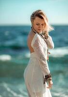 Woman beach sea white dress. The middle-aged looks good with blonde hair, boho style in a white long dress with beach decorations on the neck and arms. photo