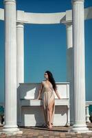 A woman in a long dress stands in front of a piano. The piano is white and has a few keys visible. The woman is posing for a photo, and the overall mood of the image is calm and serene. photo