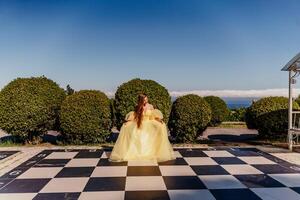 Woman yellow dress chess. A beautiful woman in a long puffy yellow dress poses on a chessboard in the park. photo