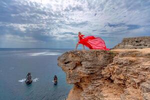 un mujer en un rojo seda vestir soportes por el océano, con montañas en el fondo, como su vestir se balancea en el brisa. foto