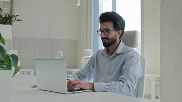 arabo indiano musulmano uomo d'affari etnico uomo Lavorando con computer a aziendale ufficio allegro contento manager dipendente programmatore digitando il computer portatile navigazione sito web attività commerciale opera guardare a telecamera Sorridi video