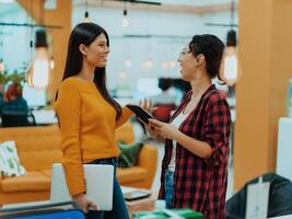 retrato de dos mujer de negocios hablando a cada otro mientras en pie en un moderno negocio oficina con su colegas utilizando ordenador portátil y tableta.. márketing concepto. multiétnico sociedad. foto