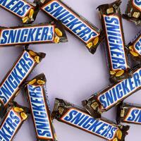 Many Snickers chocolate bars lies on pastel lilac paper. Snickers bars are produced by Mars Incorporated. Snickers was created by Franklin Clarence Mars in 1930 photo