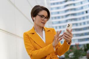 Smiling  beautiful  woman with short haircut  in yellow jacket and pants using mobyle phone. Standing over modern city. Outdoor photo. photo