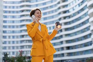 sonriente hermosa mujer con corto Corte de pelo en amarillo chaqueta y pantalones utilizando móvil teléfono , participación taza de café . en pie terminado moderno ciudad. foto