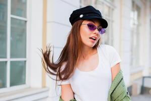 contento morena mujer posando al aire libre . elegante sombrero, Gafas de sol y verde chaqueta. foto