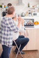 Senior couple saying hello to daughter and niece. Happy senior woman during a video conference with family using laptop in kitchen. Online call with daughter and niece. Elderly person using modern communication online internet web techonolgy. photo