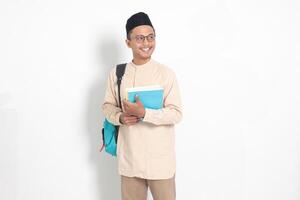 Portrait of excited student Asian muslim man in koko shirt with skullcap carrying backpack, while holding his school books. Islamic education concept. Isolated image on white background photo