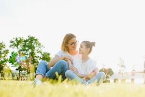 Happy and friendly family in nature. Emotional and young mother hugs her smiling little daughter lying on the grass. Summer park. Positive emotions.Childhood. Summertime.Lifestyle photo