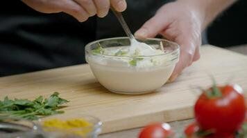 The chef mixes garlic and herbs with sour cream in a saucepan. They make a delicious white sauce, which is used in the restaurant kitchen to serve meat and fish dishes. Close up. Slow motion. 4k video