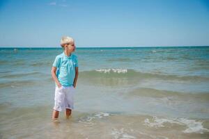 Handsome teenager boy of European appearance with blond hair in a blue T-shirt, and white shorts,stands behind blue sky in the sea water, and looks thoughtfully into the distance. Summer vocation concept.Handsome teenager boy portrait concept.Copy space. photo