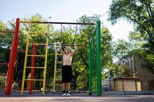 un atlético niño agarrado el anillos a hacer un levantar. calle rutina de ejercicio en un horizontal bar en el colegio parque. foto