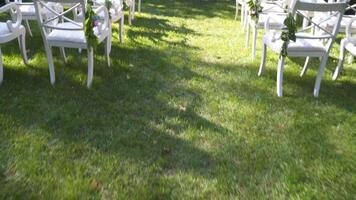 Wedding decor for outdoor cerimony. Wedding arch and chairs for guests in white color on green grass in sunny weather. Backyard wedding venue. Furniture and decoration for outside wedding party. video