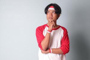 Portrait of attractive Asian man in t-shirt with red and white ribbon on head, standing against gray background, thinking about question with hand on chin photo