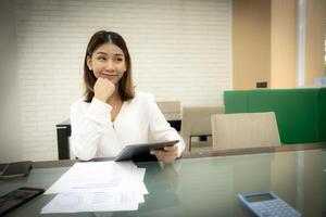 Beautiful asian business woman is sitting with a doubtful expression and holding tablet on her desk. photo