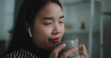 selettivo messa a fuoco, faccia di contento giovane asiatico donna godere ascoltando per il musica con senza fili cuffie mentre seduta bevanda caffè su divano nel vivente camera a casa, tempo libero e stile di vita, libero tempo video