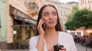 Young caucasian european woman talking chat mobile phone cellphone city outside cheerful happy girl smiling drinking holding cup coffee cafe businesswoman urban gen z street call discuss friendly talk video