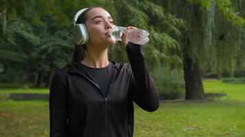 contento hermosa caucásico mujer aptitud niña sonrisa en ciudad parque deporte al aire libre hembra escuchando música auriculares relajarse bebida agua sano estilo de vida refresco sed suministro Bebiendo agua hidratación video