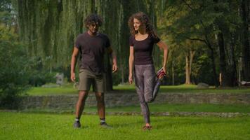 Active healthy couple Indian Arabian man and Caucasian woman athletes doing sport morning workout stretching body muscles warm-up exercises in park outdoors sporty people diverse male female training video