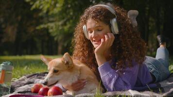 feliz retrato animal e proprietário sorridente caucasiano gen z menina ouço música dentro fones de ouvido deitado em cobertor com adorável cachorro galês corgi ao ar livre natureza jovem mulher abraço beijo pequeno cachorro dentro cidade parque final de semana video