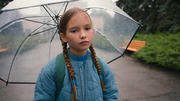 cansado pequeno menina estudante criança olhando às Câmera entediado infeliz mau clima chuva guarda-chuva cidade parque ao ar livre fadiga úmido molhado face filha criança aluno descendência solitário tristeza chuva desconforto video