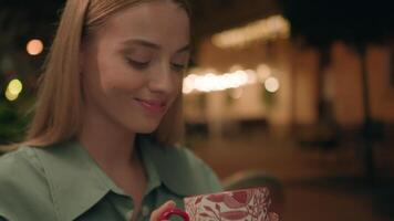 Close up young European woman blonde holding cup drink hot refreshing tasting herbal tea cozy warmth city outside nighttime cafe restaurant visitor relaxed healthy satisfaction joyful smile aroma lady video