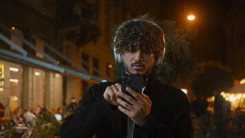 indisch Latino arabisch Mann im Kopfhörer mit Handy, Mobiltelefon Telefon Smartphone Handy Hör mal zu Musik- gehen Stadt draußen nachdenklich Kerl Geste Nacht Vergnügen heiter Gerät Technologie Lied Rhythmus Internet video