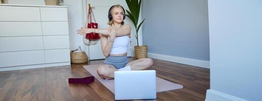 bienestar y aptitud concepto. mujer en auriculares, se sienta en yoga estera, relojes rutina de ejercicio en computadora portátil, estiramientos brazos, lo hace ejercicios foto