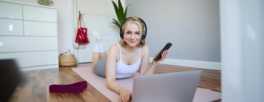 Portrait of young sporty woman, connects to online workout training session, doing exercises, lying on rubber mat, listens to fitness instructor in headphones, holding mobile phone photo