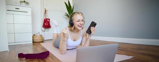 Portrait of young woman workout, watching exercise s on laptop in headphones, lying on rubber mat with mobile phone and smiling photo