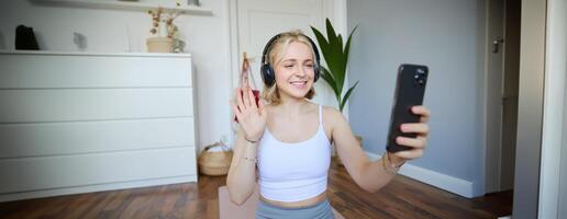 retrato de joven deportivo mujer en auriculares, registros en su teléfono inteligente, En Vivo transmisión y diciendo Hola a seguidores mientras haciendo rutina de ejercicio formación a hogar foto