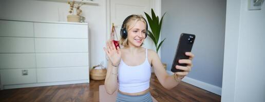 Portrait of young woman with smartphone and headphones, waving hand at mobile phone camera, live streaming during workout photo