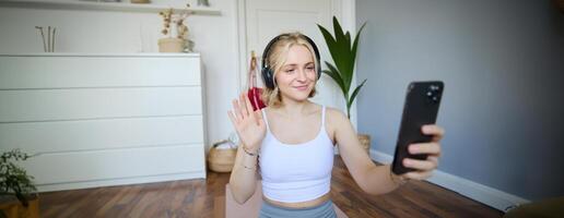 Portrait of woman waving hand at smartphone, wearing wireless headphones, working out and recording for social media account about fitness photo