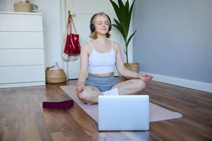 retrato de concentrado mujer en auriculares, meditando, escuchando a calma música y práctica yoga, seguir instrucciones en computadora portátil, sentado en caucho estera foto