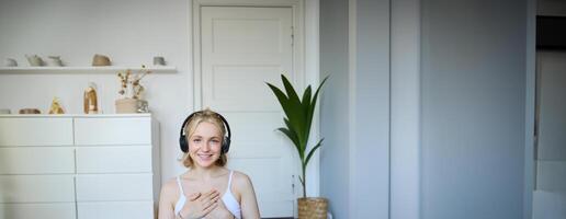 Portrait of woman feeling relaxed and in peace after meditation or yoga training at home, holding hands on chest, wearing headphones, smiling at camera photo